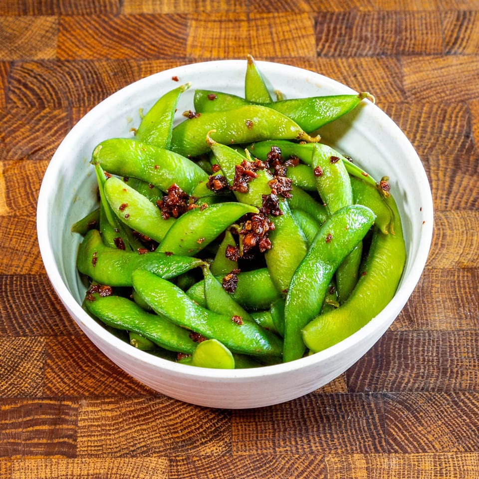 A white bowl filled with vibrant green edamame pods, topped with a savory brown seasoning.