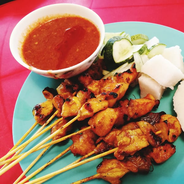 Grilled chicken satay skewers with peanut sauce, cucumber, and rice cakes on a blue plate.